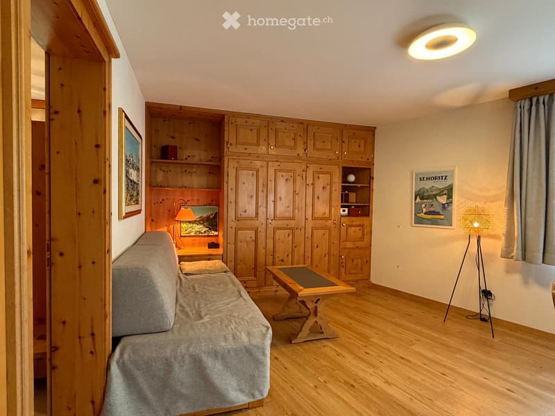 Cozy, furnished room with wooden floors, a sofa, a small table, a lamp, and built-in wooden cabinets.