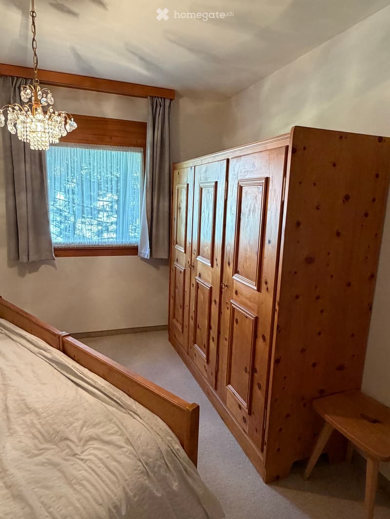 Cozy bedroom with a wooden bed, large wardrobe, small wooden stool, and a chandelier hanging from the ceiling. The room features a large window with curtains and a view of trees outside.