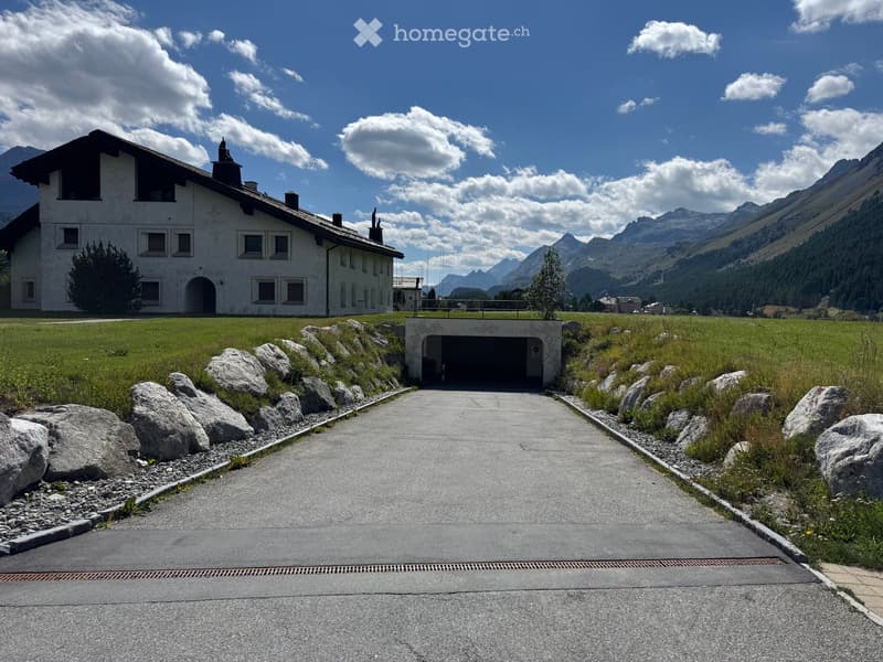 A large white building with multiple windows and a chimney, situated on a grassy area with a paved pathway leading to a garage entrance, surrounded by mountains.