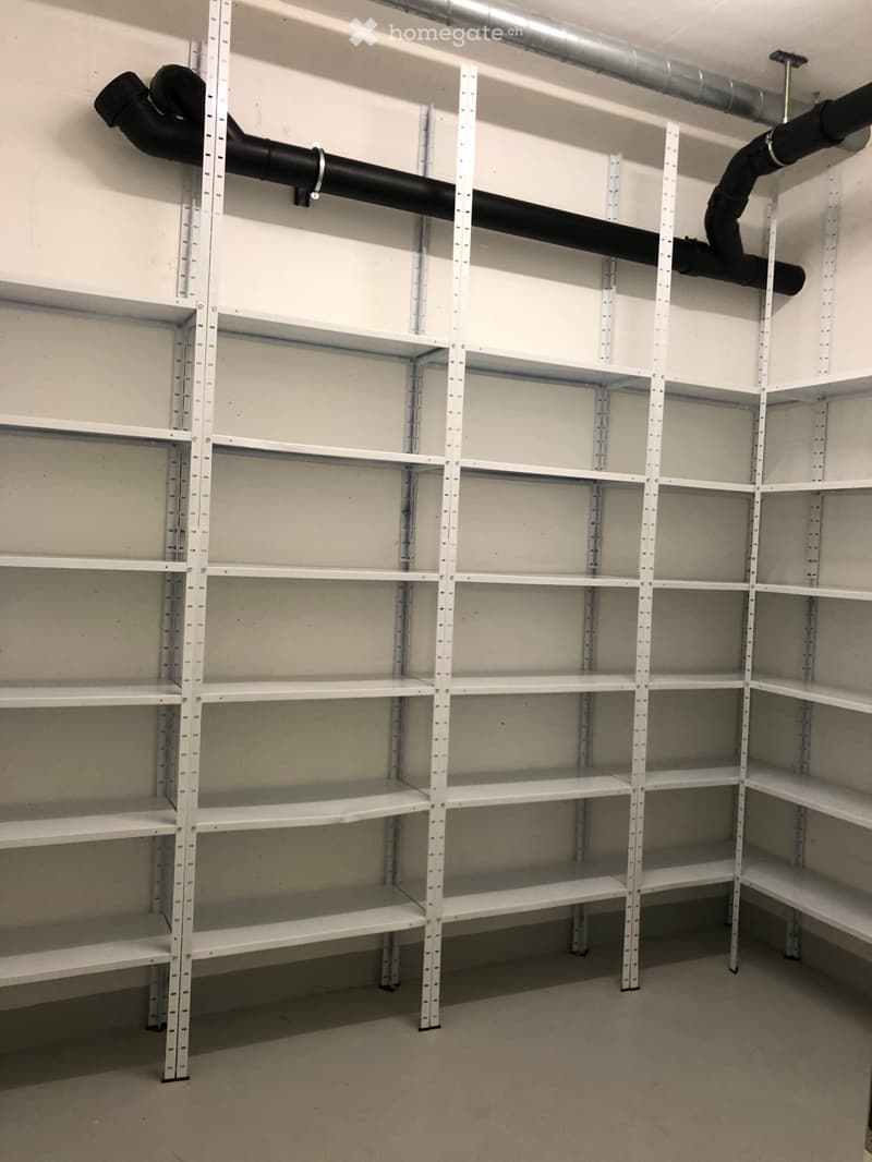 Empty storage room with white metal shelving units and black ventilation pipes running along the ceiling.