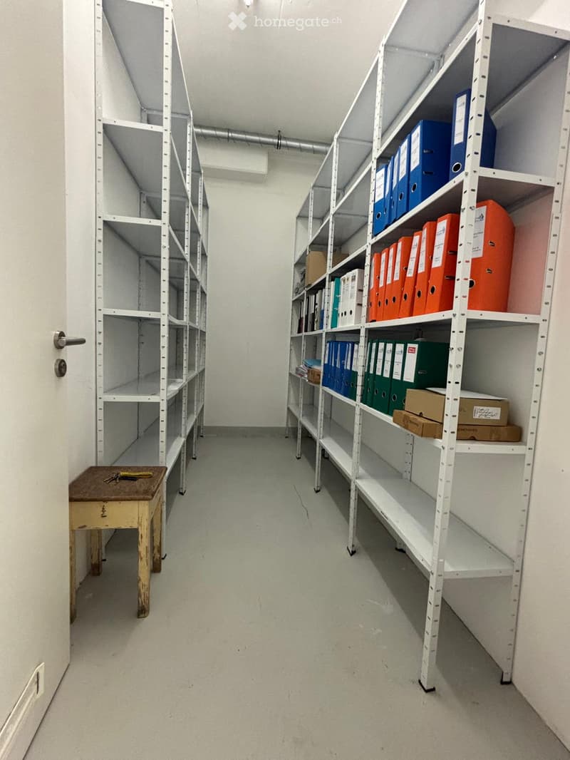 A storage room with metal shelving units filled with folders and boxes, a small wooden stool with keys and tools on it, and a door on the left side.