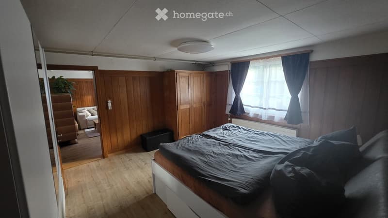 Cozy bedroom with wooden walls, a large bed with dark bedding, a wardrobe, a small living area with a sofa and table, and a plant near the doorway.