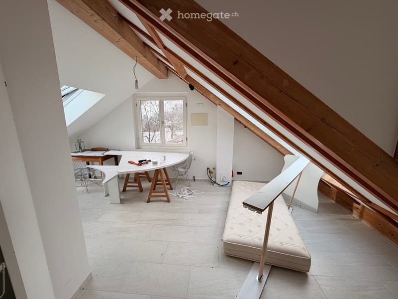 Bright attic room with exposed wooden beams, white walls, and large windows. Contains a table with chairs, a bench, and a lamp.