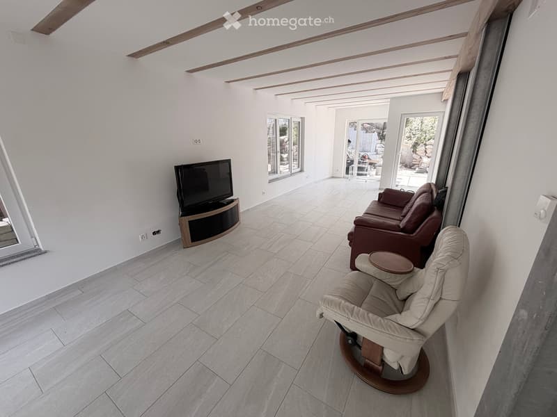 Modern living room with white walls, tiled floor, large windows, and sliding glass doors leading to an outdoor area. Features a brown leather sofa, a beige armchair, a small round table, and a flat-screen TV on a wooden stand.