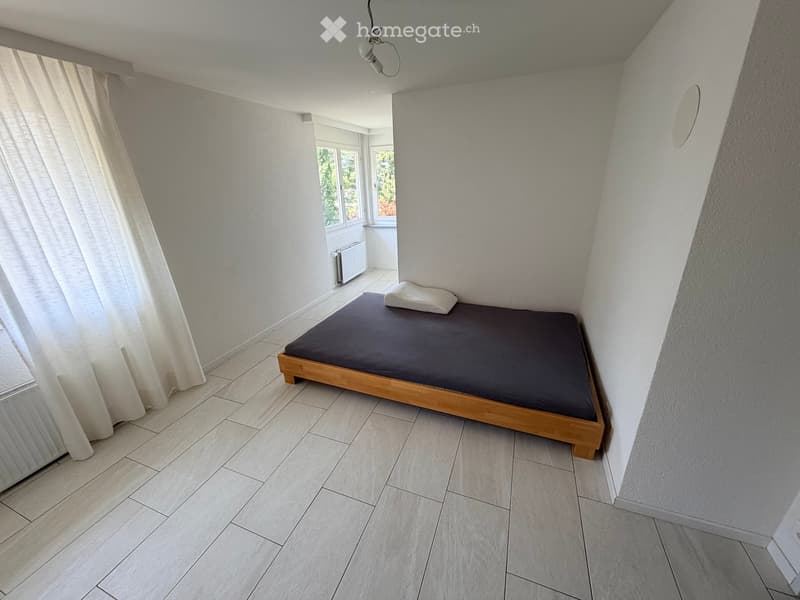 Minimalist bedroom with a single wooden bed, dark grey mattress, and a single white pillow. Light grey walls, tiled floor, and large windows with white curtains.