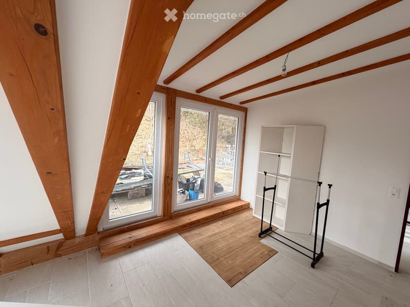 An attic room with wooden beams, large windows, and a wooden floor. The room is empty, with no furniture or furnishings.