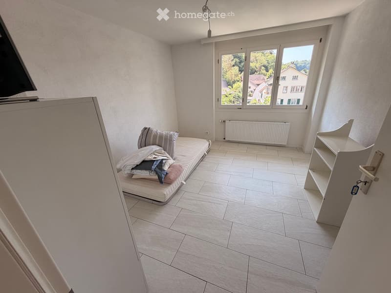 Small furnished room with tiled floor, bed with pillows and blankets, radiator, shelf, and large window with view of greenery.