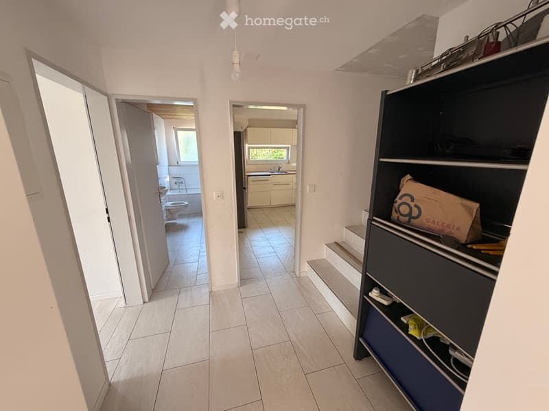 Bright hallway with tiled flooring, white walls, and open doors leading to a bathroom and kitchen area. A built-in storage unit with shelves is visible on the right side.