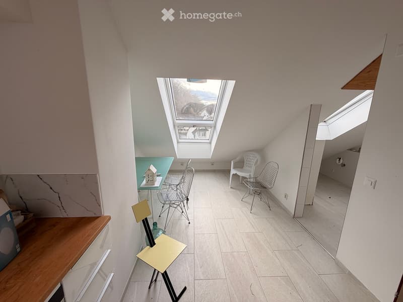 Bright, airy room with white tiled flooring, white walls, and skylight windows. Contains a small table with a decorative house model, several chairs, and a stool.