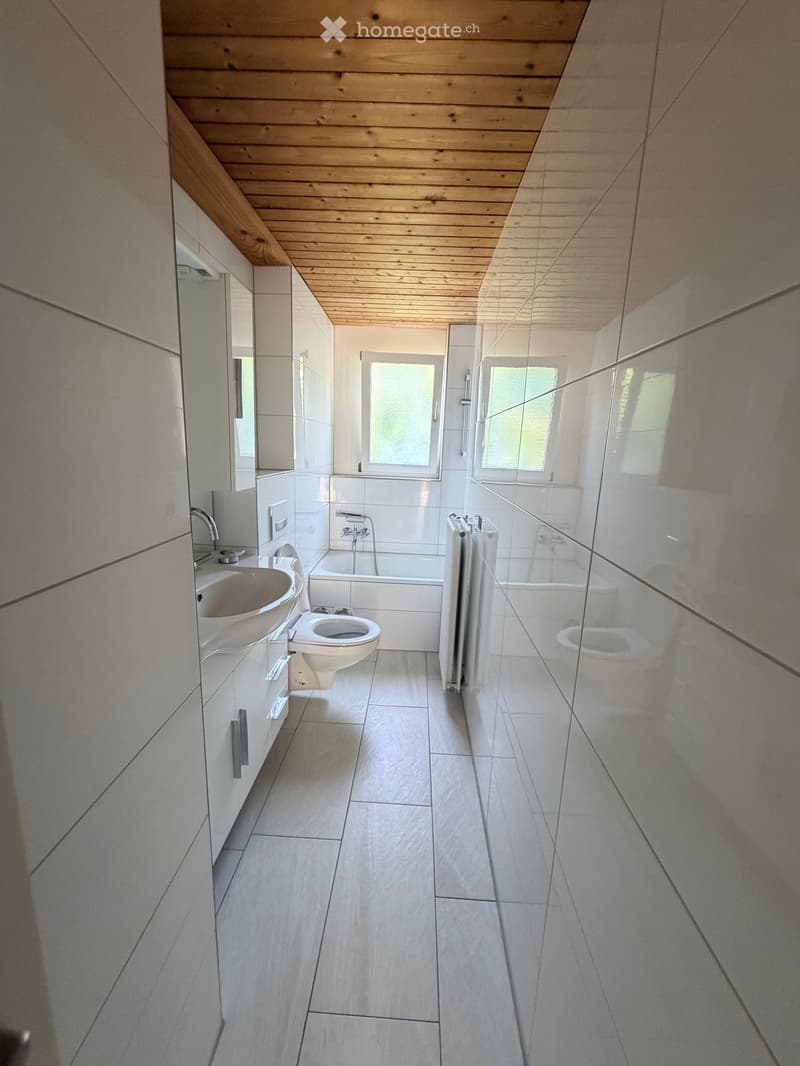 Modern bathroom with a wooden slatted ceiling, white tiled walls and floor, a sink with a mirror, a toilet, a bathtub, and a glass-enclosed shower area.