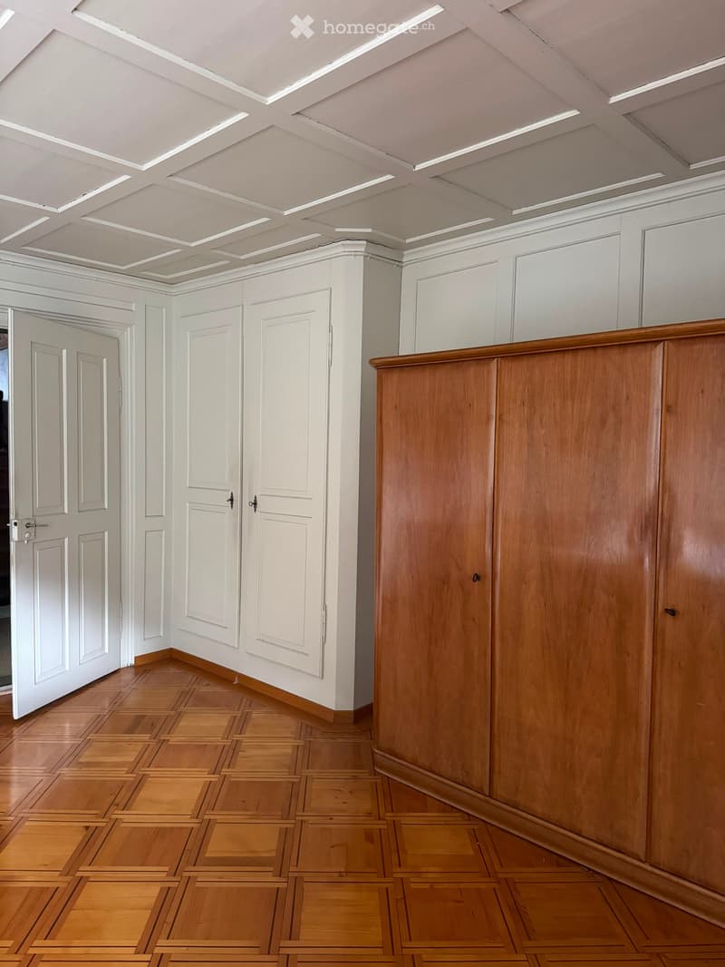 Empty hallway with wooden flooring, white walls, and multiple white doors. The ceiling features recessed lighting and a decorative pattern.