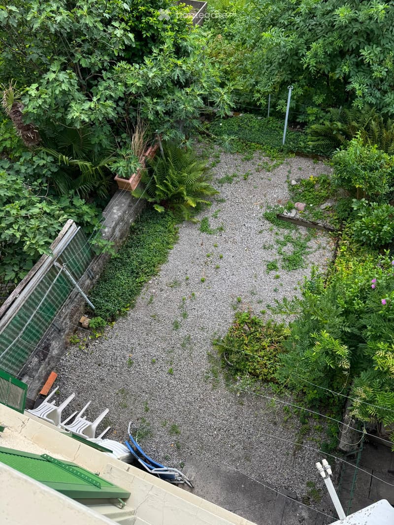 A small, lush garden area with gravel ground, various plants, and a few outdoor chairs.