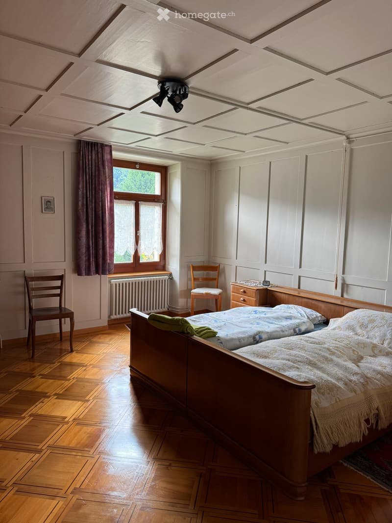 Cozy bedroom with wooden floor, large window with curtains, bed with patterned bedding, two chairs, nightstand, radiator, and a picture frame on the wall.