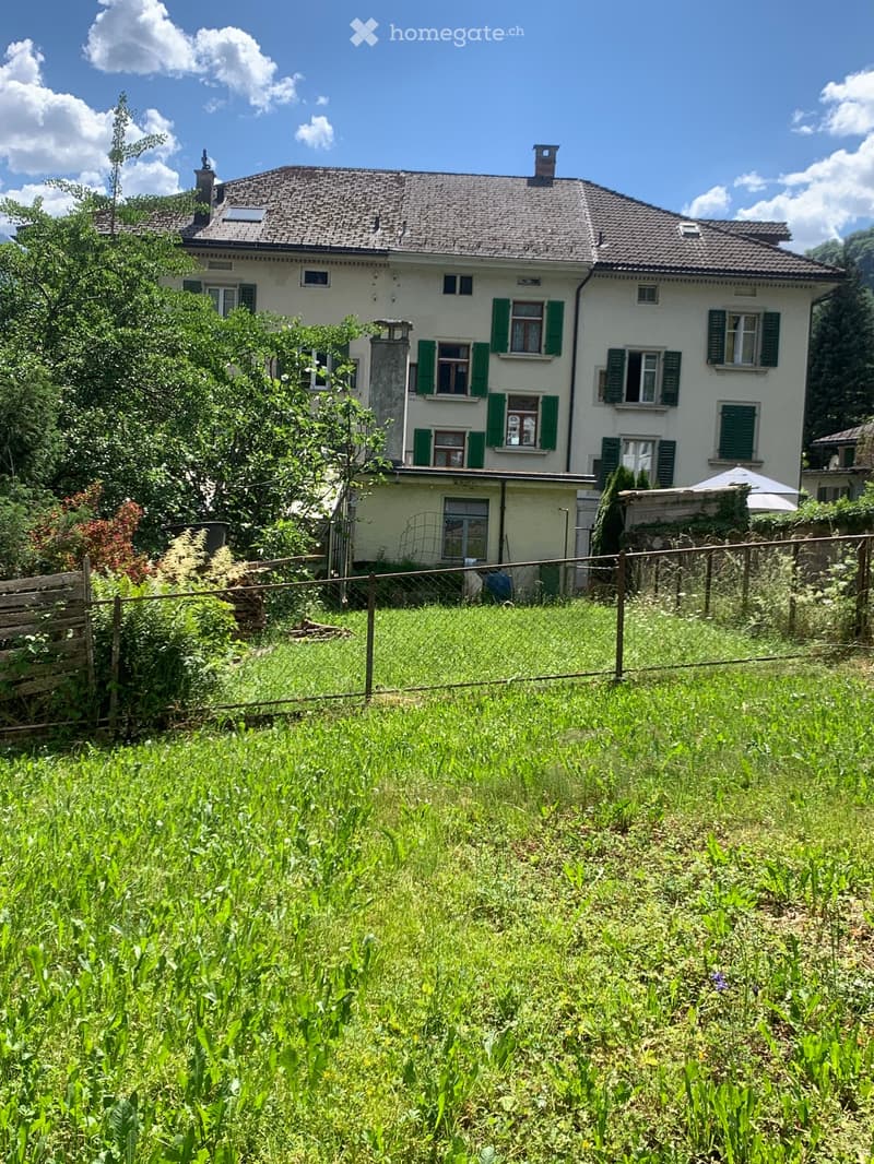 A two-story white house with green shutters, surrounded by a grassy yard and trees.