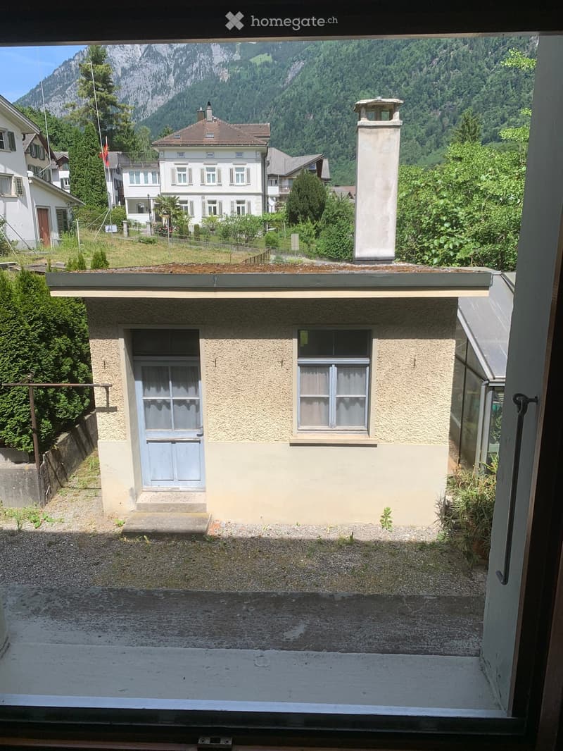 Balcony overlooking a small building with a door and window, surrounded by greenery and distant mountains.