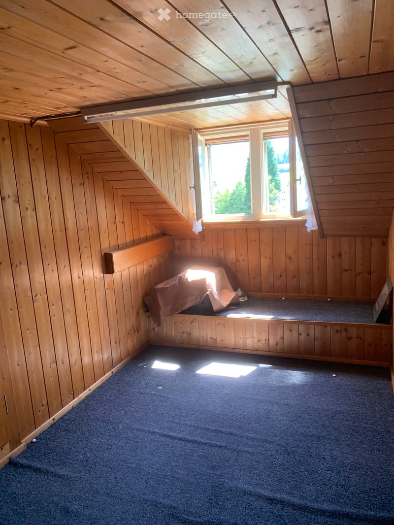 An attic room with wooden walls and ceiling, carpeted floor, and two windows letting in natural light. The room is empty with no furniture.
