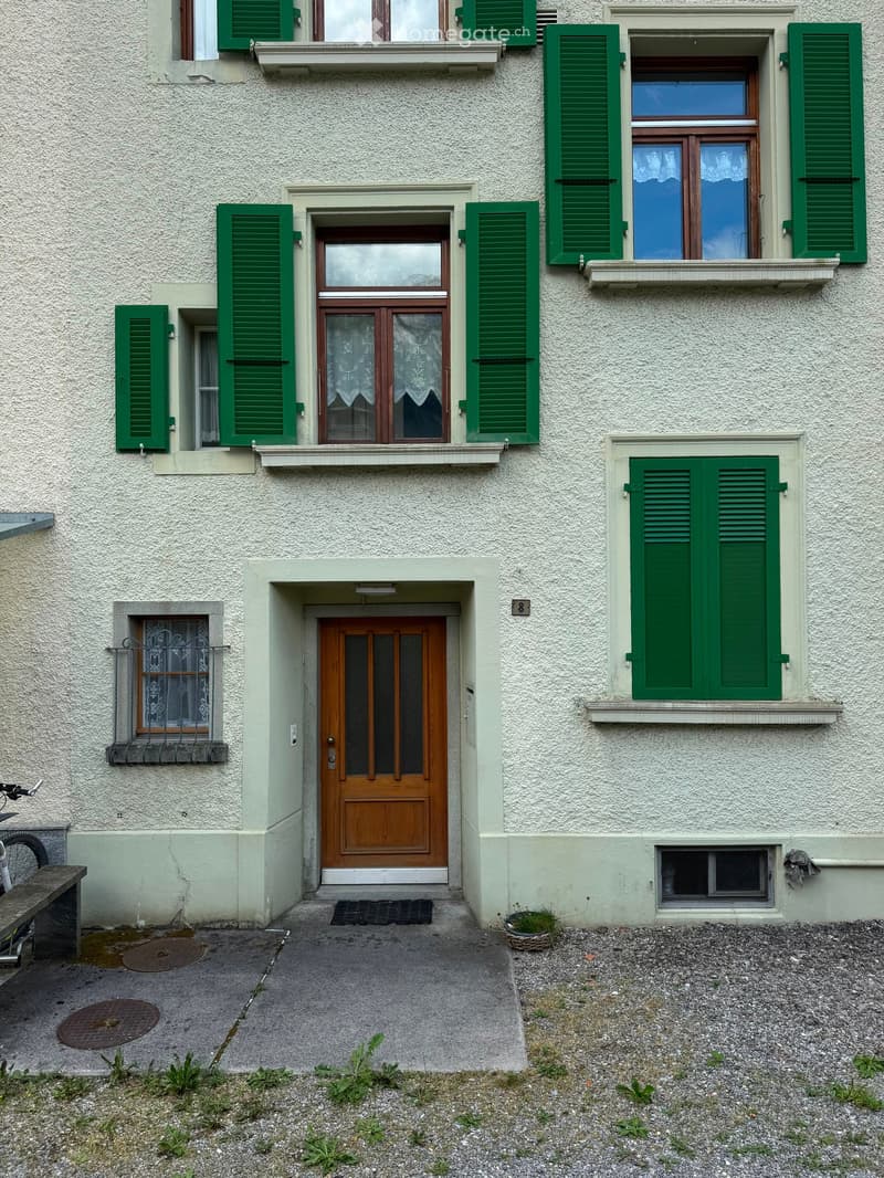 Two-story residential building with light-colored textured walls, green shutters, and multiple windows. Features a central wooden front door with glass panels, a small doormat, and a small potted plant near the entrance.