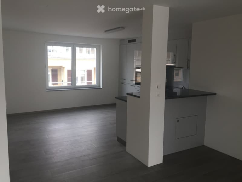 Empty room with wooden flooring, white walls, and a large window with a view of a building outside. There is a kitchen area with cabinets and a countertop, but no furniture or appliances are present.
