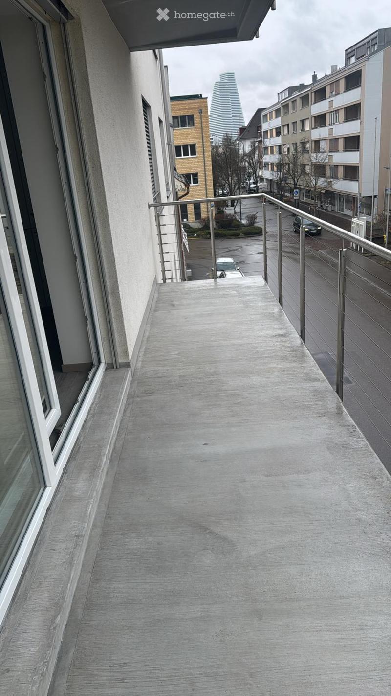 Balcony with metal railing, concrete floor, overlooking a city street with buildings and parked cars.