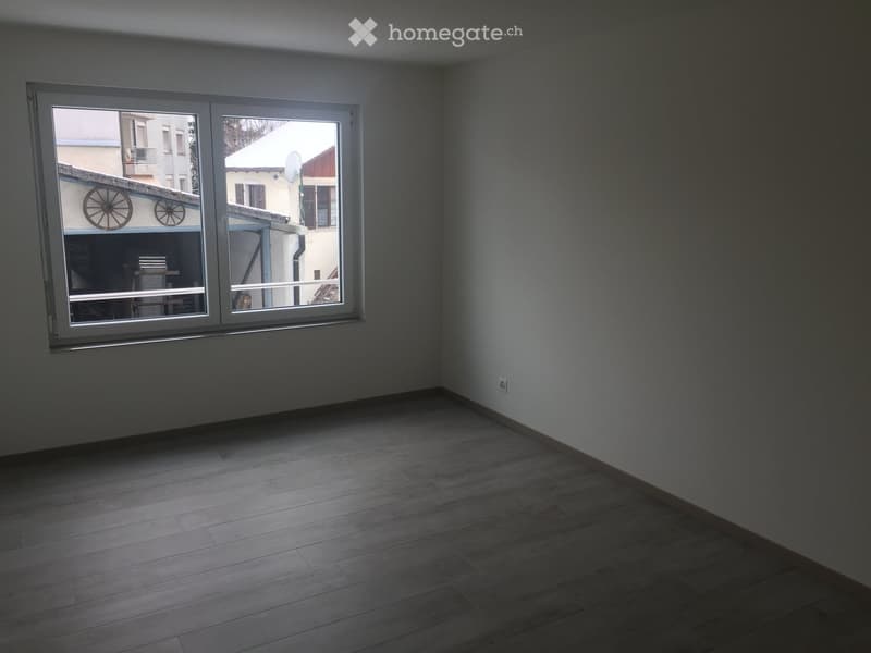 Empty room with wooden flooring and large windows, showing a view of the outside area.