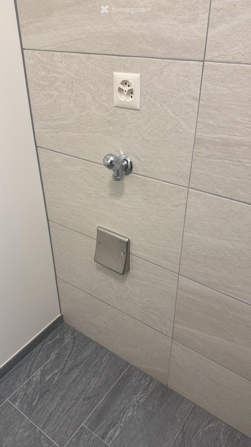 Walls covered with large rectangular tiles, floor covered with smaller rectangular tiles, electric outlet and pipe fitting on the wall