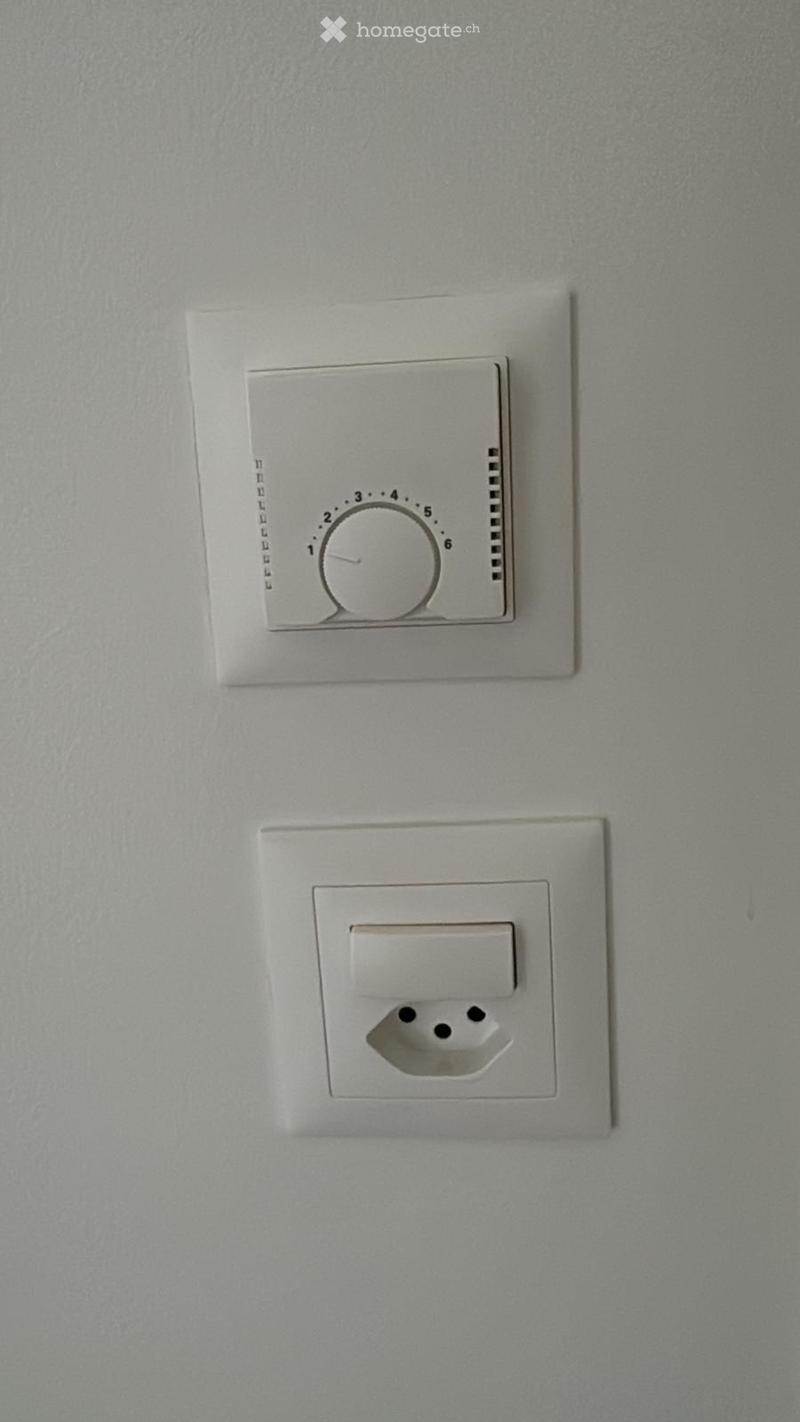 A plain wall with a thermostat and a power outlet, no furniture or furnishings present.
