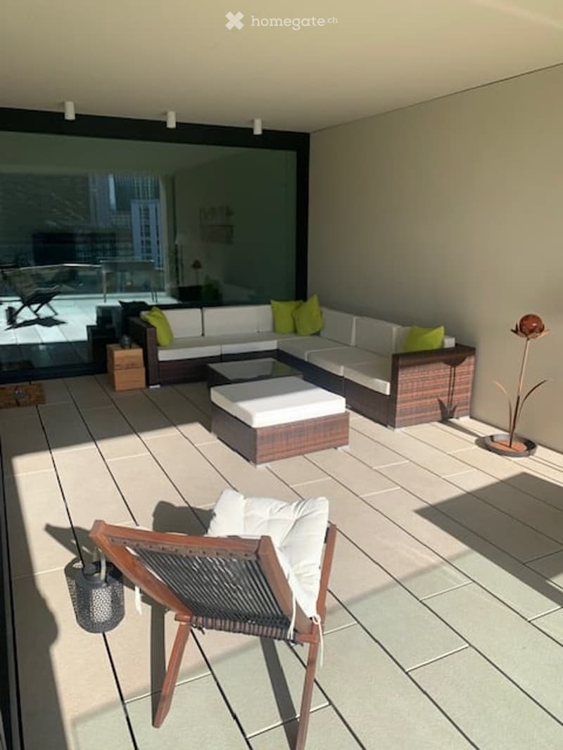 Modern balcony with large glass windows, white sectional sofa, green cushions, wooden coffee table, and a single wooden chair.