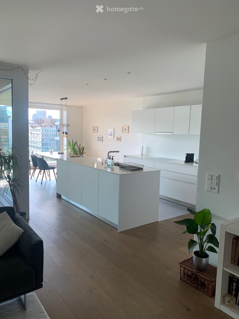 Modern open-plan living and dining area with a kitchen island, white cabinets, a sofa, a coffee table, and a dining table with chairs. The space features wooden flooring, large windows with a city view, and decorative plants.