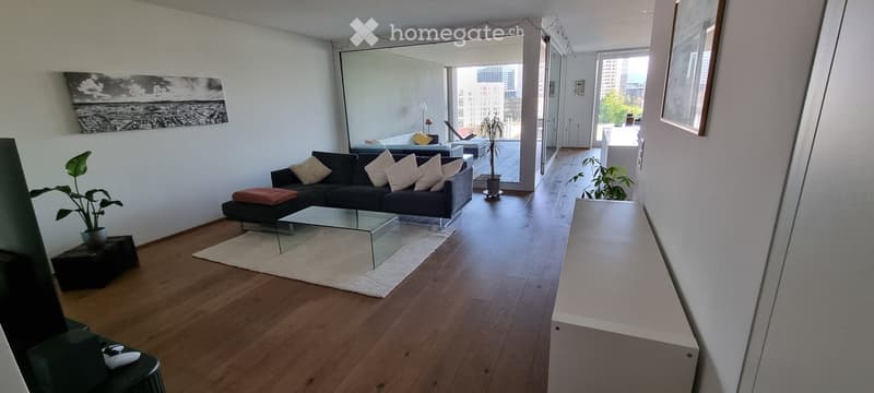 Modern living room with wooden flooring, large windows, a black sofa with white and red cushions, a glass coffee table, side tables with plants, and a mirror on the wall.