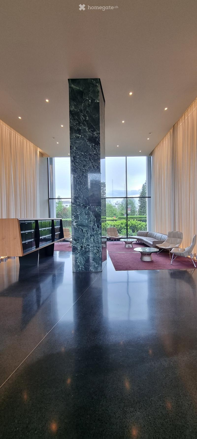 Modern lobby area with a black polished floor, a large central marble pillar, and a seating area with a sofa, chairs, and a glass coffee table.