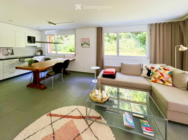Bright, open-plan living and dining area with a beige sofa, glass coffee table, wooden dining table, and modern kitchen. Features large windows with curtains, a floor lamp, and decorative items on the coffee table.