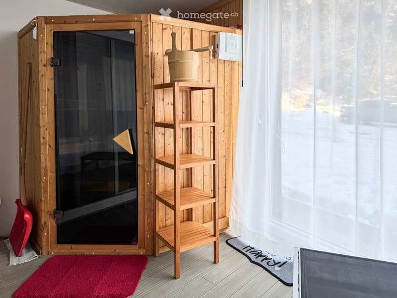 A wooden sauna with a glass door, a wooden shelf holding a bucket, and two rugs on the floor.