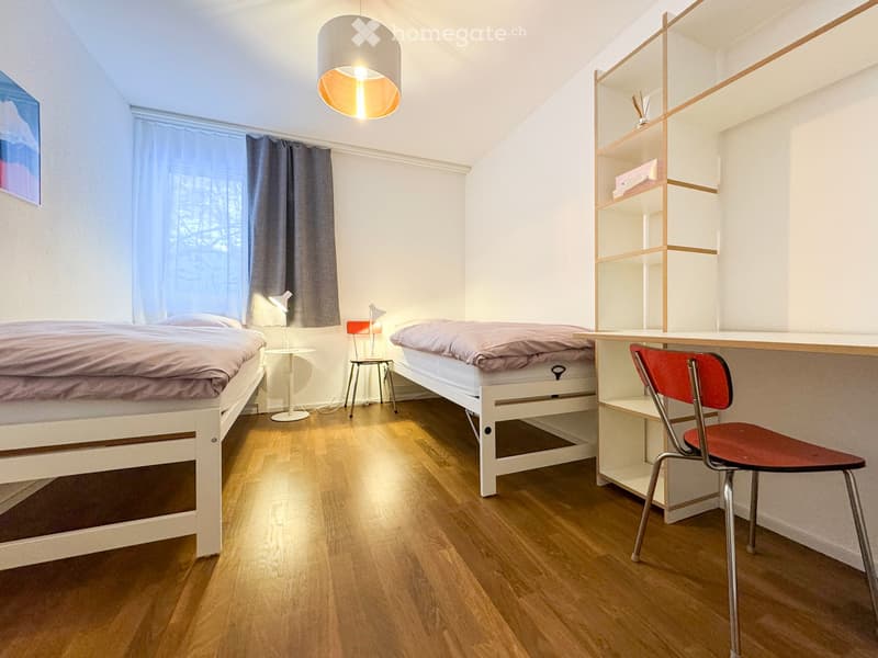 Cozy bedroom with two single beds, a desk, a chair, and a shelf. The room features wooden flooring and large windows with curtains.