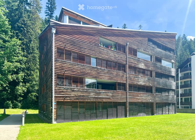 Modern multi-story building with wooden facade, large windows, and balconies, surrounded by greenery.