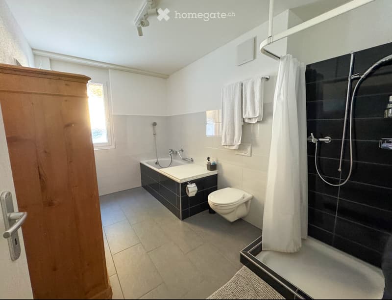 Modern bathroom with a bathtub, toilet, shower, towel rack, and a wooden cabinet. The space features white tiles, a large window, and a sleek design.