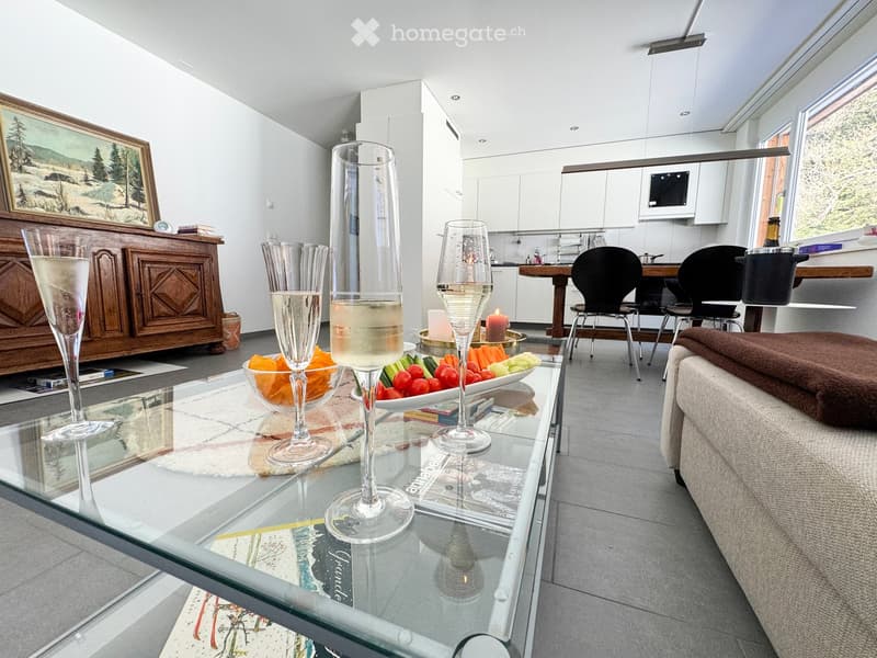 Modern living room with a glass coffee table, sofa, chairs, and a small kitchen area with cabinets and a microwave. The room features a painting on the wall and a bowl of snacks on the coffee table.
