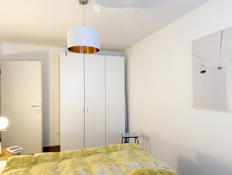 Modern bedroom with a large bed, white wardrobes, a hanging pendant light, a small stool, and a mirror on the wall.