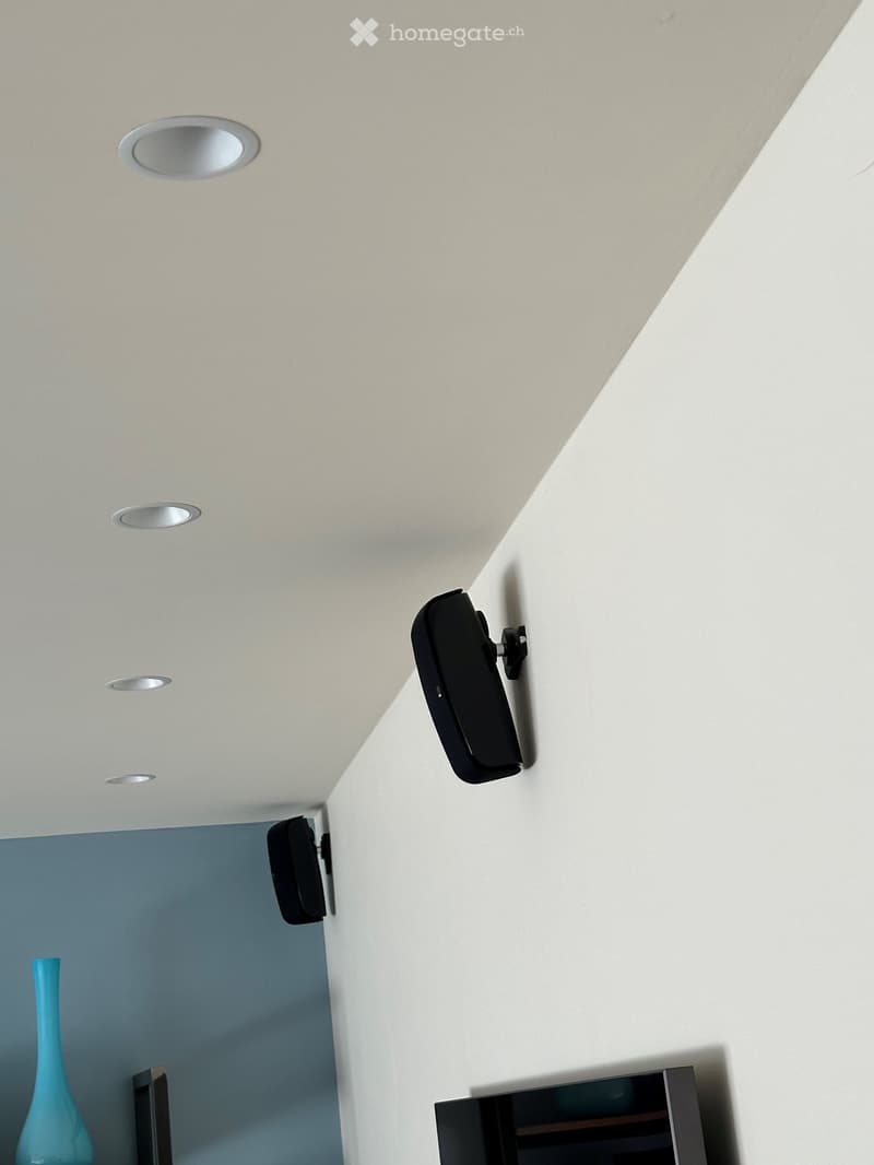 Modern living room with a flat-screen TV mounted on the wall, two wall-mounted speakers, recessed ceiling lights, and a blue vase on a shelf.