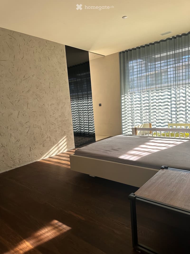 Modern bedroom with wooden flooring, large window with sheer curtains, bed with dark bedding, nightstand, and chair visible.