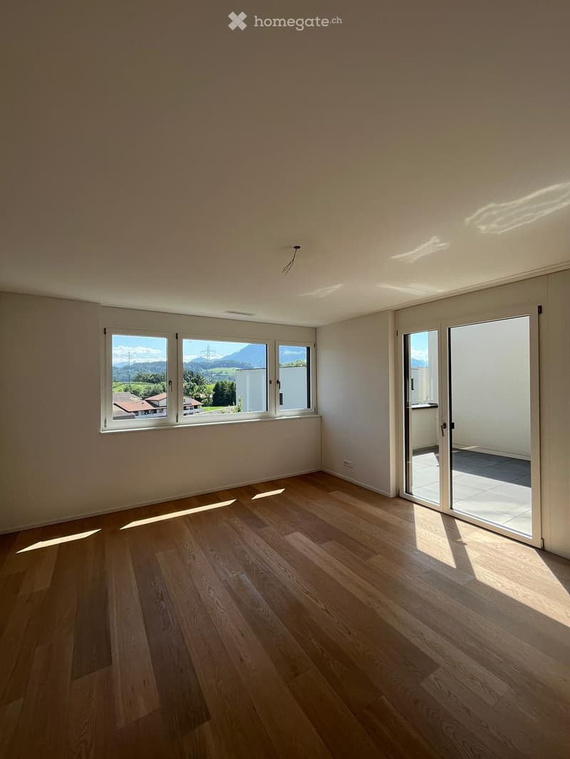Empty room with light wooden flooring, large windows, and sliding glass doors leading to a balcony.