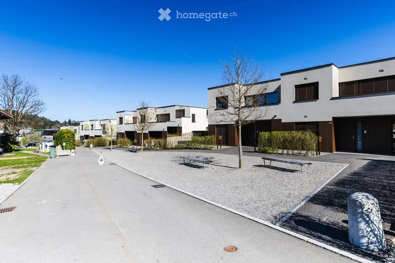 Row of modern houses, white exterior, brown accents, large windows, gravel parking area, benches, trees, clear blue sky