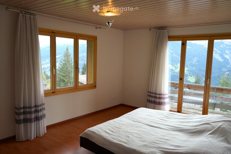 Bright bedroom with wooden floors, large windows and sliding doors offering mountain views, a neatly made bed, and light-colored curtains.
