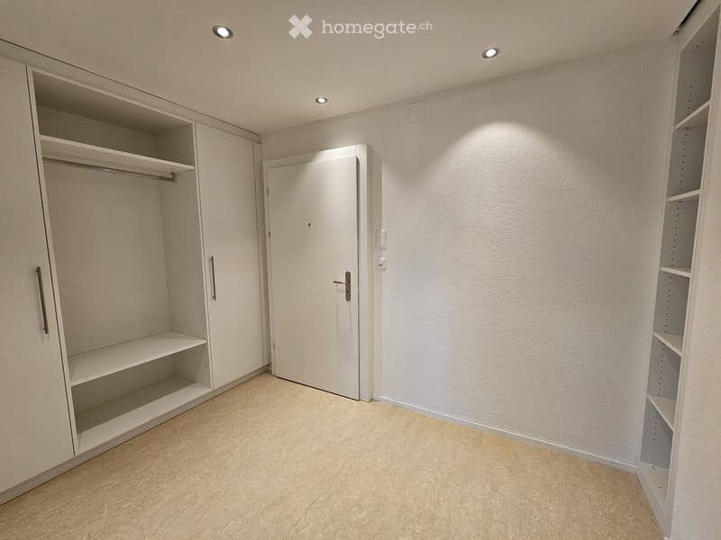 The image shows a bedroom with a white door, a built-in closet, and shelving units on the walls. The room has a light-colored floor and is well-lit with recessed lighting.