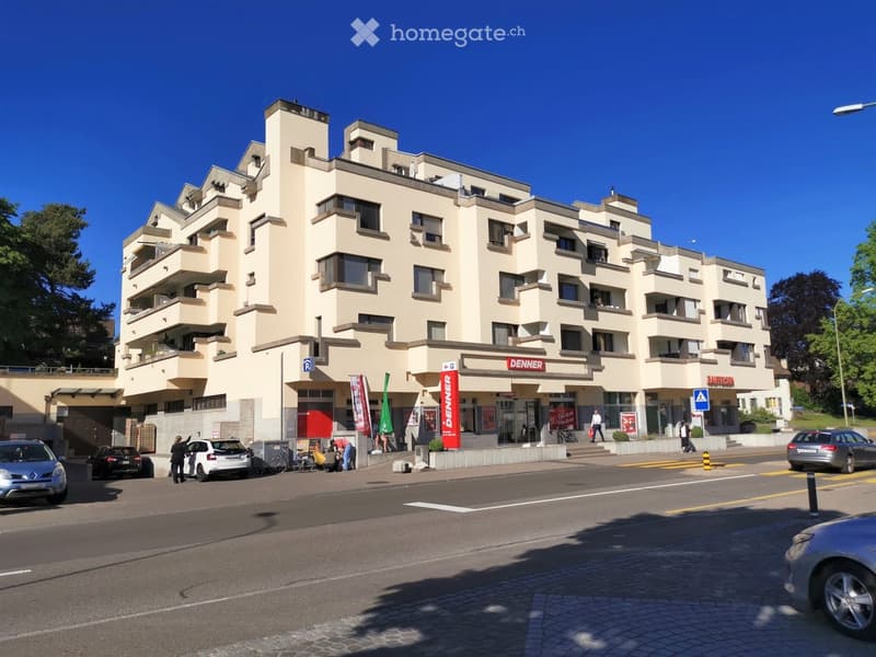 Multi-story apartment building with a modern, angular design. The building has numerous balconies and terraces, and there are several commercial spaces on the ground floor, including a Denner store.