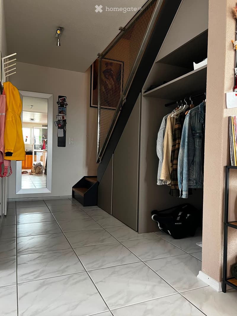 Hallway with tiled floor, staircase leading upwards, coat rack with jackets, open closet with hanging clothes and a stroller, and a mirror on the wall.