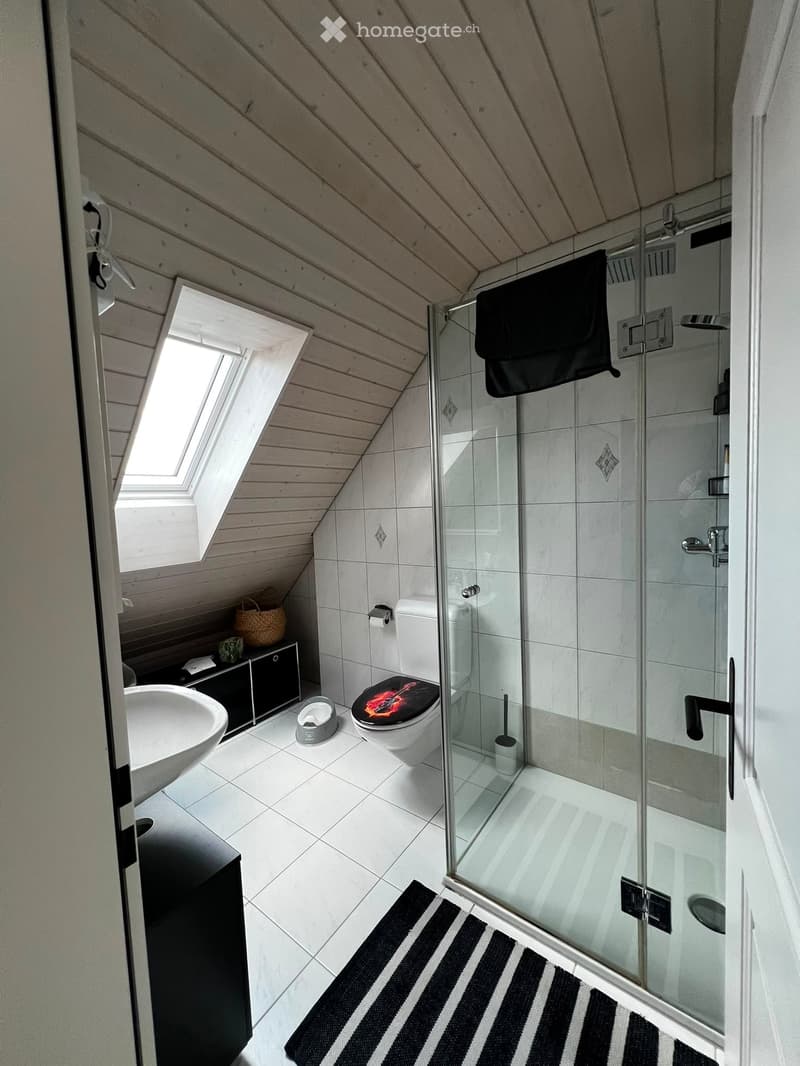 Modern bathroom with white tiles, a glass-enclosed shower, a sink, a toilet with a decorative seat cover, a small black cabinet, and a wicker basket on the cabinet.