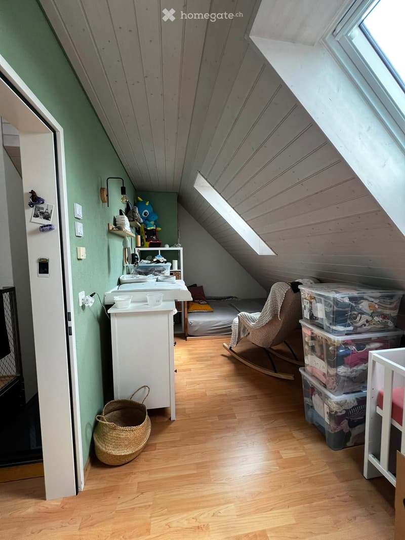 Cozy attic bedroom with wooden floors, slanted ceilings, and large windows. Features a bed, rocking chair, small table, storage bins, and decorative items.