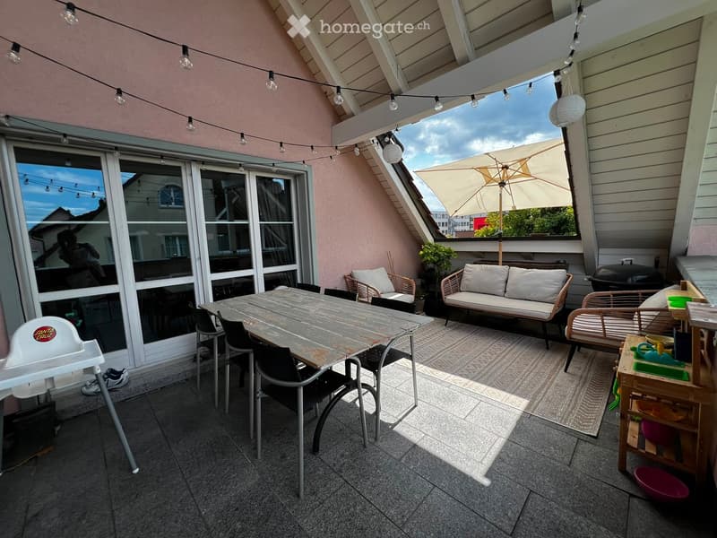 A spacious balcony with a dining table, chairs, a sofa, an umbrella, and a baby chair, offering a view of the city.