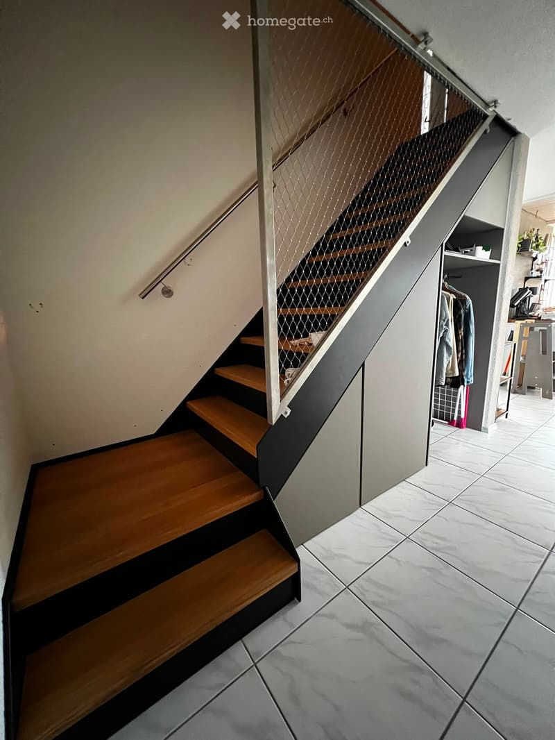 Modern hallway with wooden staircase featuring a metal mesh railing, light grey tiled floor, and adjacent closet with hanging clothes and storage items.