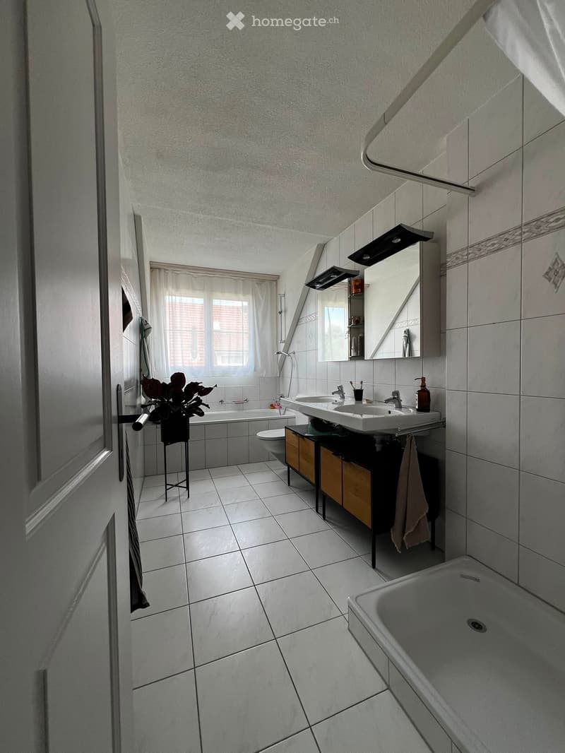 Modern bathroom with white tiles, a bathtub, a double sink, a toilet, a mirror, a small cabinet, and a towel hanging on the side.